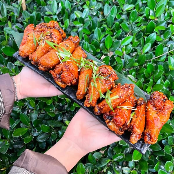 Hands holding a black platter of Kanpai's glazed chicken wings, garnished with green onions, against a leafy green background.