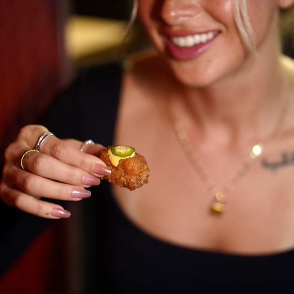 A smiling person holds up a crispy fried sushi bite topped with sauce and a jalapeño slice.