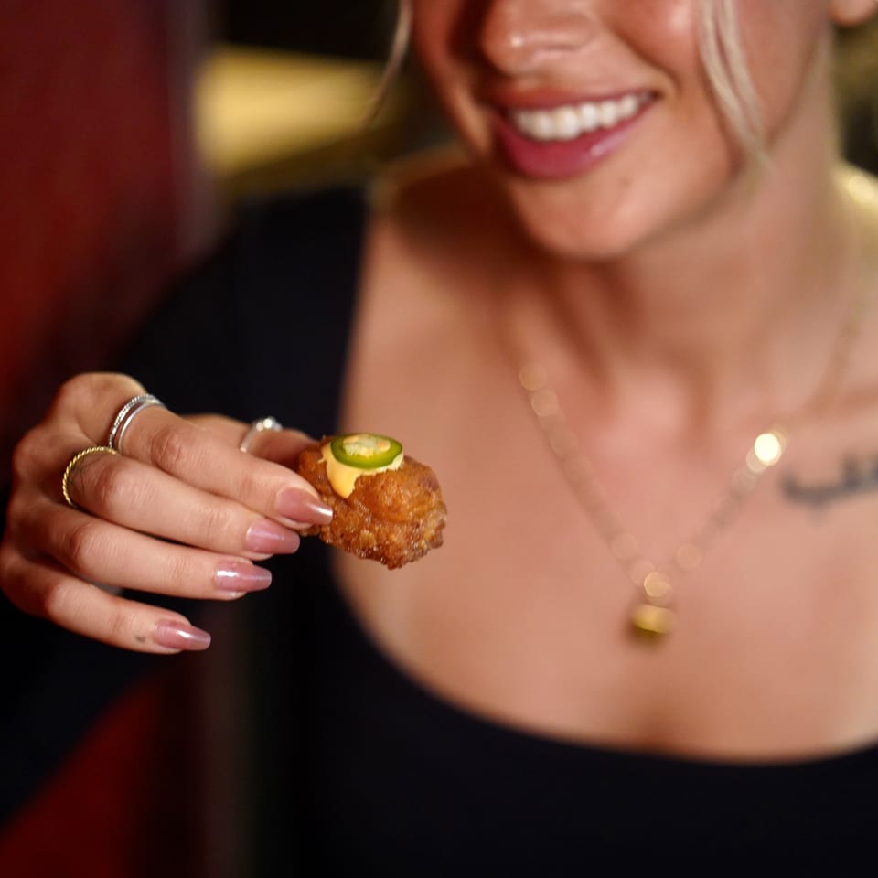 A smiling person holds up a crispy fried sushi bite topped with sauce and a jalapeño slice.