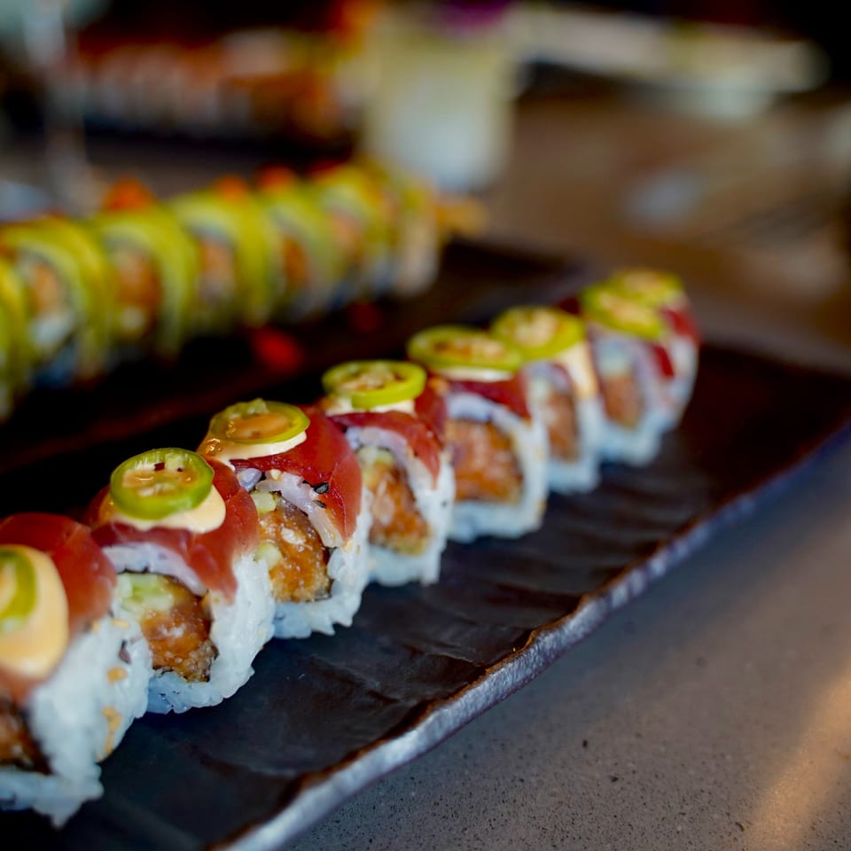 Two plates of Kanpai sushi rolls, one topped with tuna and jalapeños, the other with avocado.