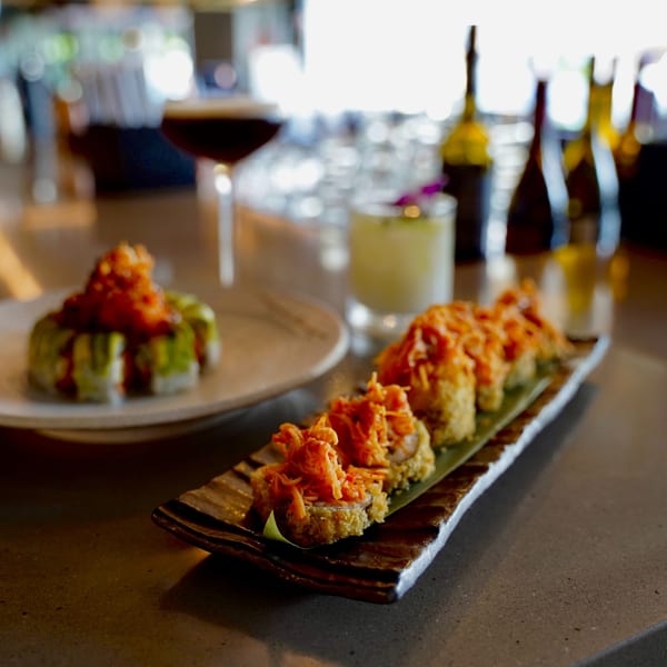 Kanpai sushi rolls, one topped with avocado and sauce, another with crispy orange topping, on a bar.